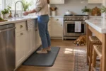Woman standing on a cushioned anti-fatigue kitchen mat by the sink in a bright modern kitchen with a dog resting nearby.